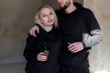 İki genç şık insan adam ve bir moda siyah sweatshirt bir kız ayakta ve elinde kahve siyah bir fincan tutarak. Oldukça güzel seksi kız içecekler kahve. Şık çift severler.