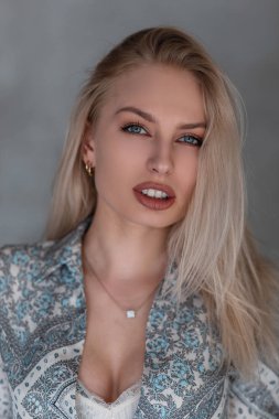 Portrait of a beautiful young blonde woman with natural make-up with blue eyes with sexy lips in a vintage shirt with a pattern in the studio against a gray wall. Attractive girl fashion model.