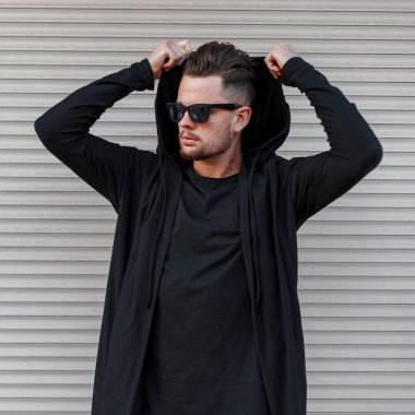 Modern metal duvar yakın bir kaput giyen siyah moda Sweatshirt Siyah bir tişört içinde koyu güneş gözlüğü şık bir saç modeli ile bir sakal ile şık genç hipster adam. Serin adam.