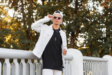 Şık bir kot tişört içinde güneş gözlüğü bir yaz beyaz ceket modern genç hipster adam bir metal çit yakın bir parkta pozlar. Güzel adam model hafta sonu zevk. Yaz Gençlik tarzı.