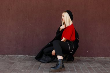 Eski bir duvara yakın oturan poz çizmeler kırmızı bir tişört uzun bir moda sonbahar ceket içinde siyah bir bere seksi kırmızı dudaklar ile modern güzel güzel kadın sarışın. Zarif kız açık havada istirahat.