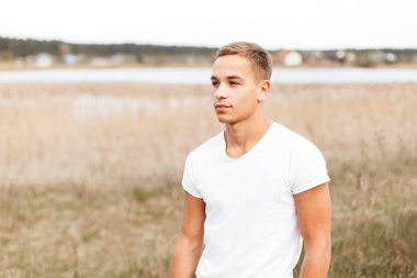 Bir yaz moda beyaz T-shirt şık bir saç modeli ile yakışıklı şık adam kuru sonbahar çim arasında bir alanda duruyor. Doğada rahatlatıcı atletik güzel adam.