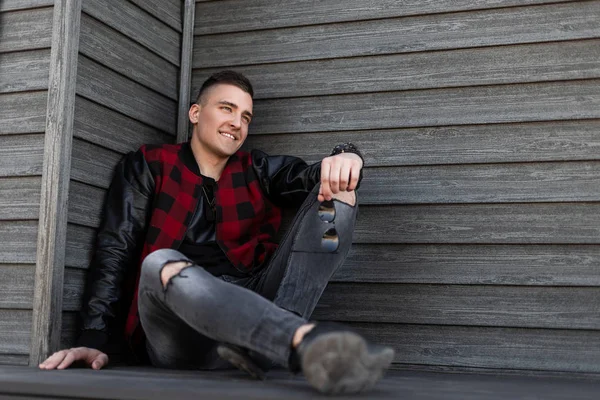 Feliz joven americano con estilo en una chaqueta a cuadros de moda con mangas de cuero en vaqueros con estilo rasgado en zapatillas de deporte relajado sentado cerca de un edificio de madera vintage. Alegre chico modelo . — Foto de Stock