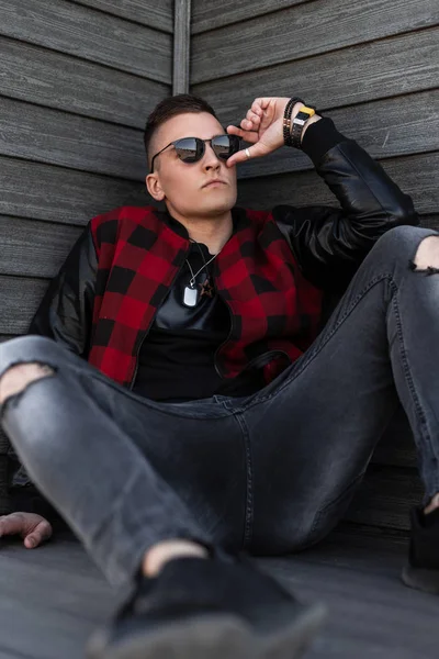 Hezký mladý hipíků v černých slunečních brýlích v módním červeném kabátku v teniskách v šedých roztrhaných džínách sedí venku u starobylé dřevěné zdi. Americký chlápek rád Odpočívej. — Stock fotografie