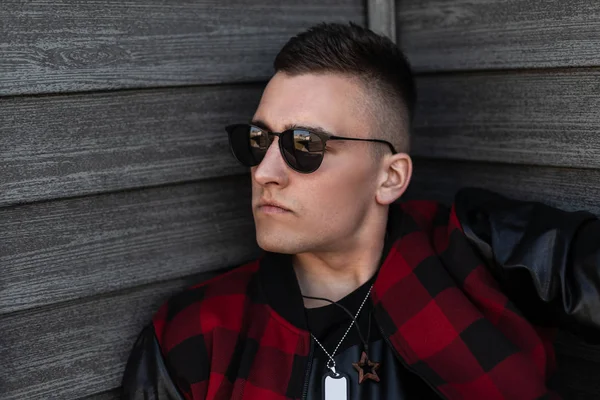 Ahşap bir duvara yakın deri kollu şık bir kırmızı ve siyah damalı ceket şık bir saç modeli ile şık siyah gözlük çekici genç hipster adam. Bir adamın yüzünün portresi. 