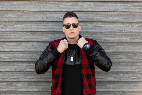 Beau jeune homme hipster dans une veste à carreaux à la mode dans un t-shirt noir avec des lunettes de soleil élégantes pose à côté d'un mur vintage en bois. Charmant mannequin moderne. Style de rue . — Photo