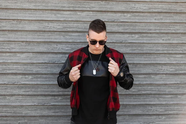 Handsome young hipster man in a stylish plaid jacket in a trendy black t-shirt with black sunglasses is posing near to a wooden vintage wall. Nice guy fashion model. American street style. — Stock Photo, Image