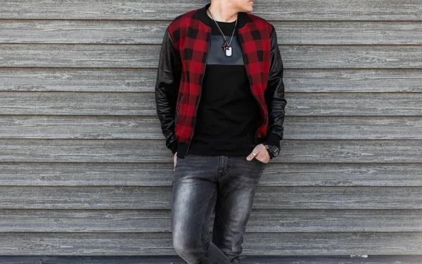 Μοντέρνος νέος άντρας με ένα μοντέρνο κόκκινο καρό μπουφάν σε ένα σκισμένο γκρίζο τζιν σε ένα μαύρο μπλουζάκι με ένα φυλαχτό να ποζάρει στο δρόμο κοντά σε έναν ξύλινο τοίχο. Σύγχρονος τύπος έξω. Κοντινό σετ ενδυμάτων ανδρών — Φωτογραφία Αρχείου