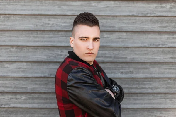 Guapo joven serio con ojos marrones en una chaqueta a cuadros rojo de moda con un peinado de moda en una camiseta con un reloj posando cerca de una pared de madera vintage. Lindo modelo de hombre. Moda masculina . — Foto de Stock