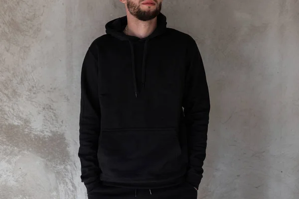 Fashionable young man with a beard in black stylish clothes is standing indoors near a concrete vintage wall. Trendy cool guy. Modern men's fashion. Close-up. — Stock Photo, Image