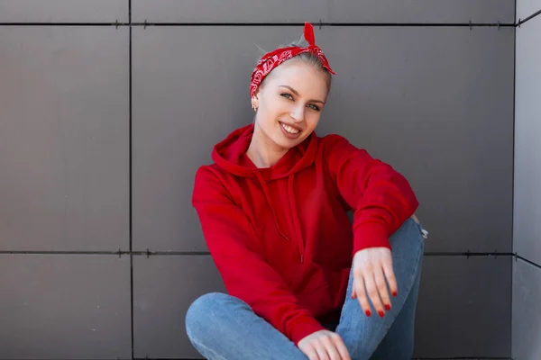 Positiv gestylte junge Frau mit Frisur in rotem Kopftuch im modischen roten Sweatshirt in Vintage-Jeans sitzt neben einem grauen modernen Gebäude und lächelt. schöne amerikanische Mädchen genießt Sommertag. — Stockfoto