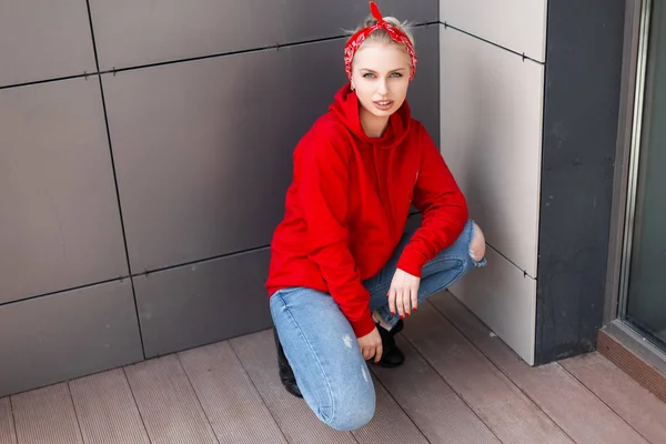 Ung modern snygg kvinna i svarta stövlar i en röd tröja i rippade jeans med en fashionabel bandana sittande utomhus nära en grå vägg. Vacker flicka åtnjuter resten. Ungdom amerikansk stil. — Stockfoto