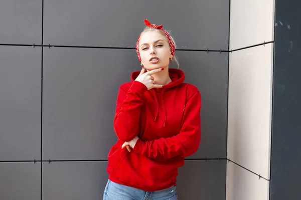 Pretty Sexy ung kvinna med blont hår i en fashionabel röd tröja med huva i en snygg bandana i vintage jeans utgör i en modern byggnad på en sommar dag. Vacker flicka. Amerikansk stil. — Stockfoto