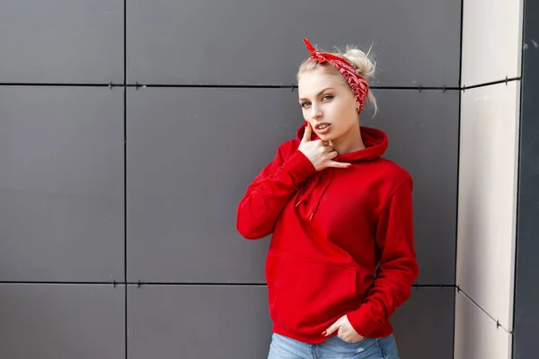 Чувственная сексуальная молодая блондинка в винтажной бандане в красной стильной толстовке в синих джинсах, позирующая на открытом воздухе возле серой стены в летний день. Американская красивая модель наслаждается отдыхом . — стоковое фото