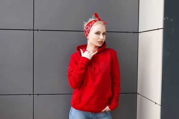 Urbain moderne jolie jeune femme dans un sweat-shirt rouge à la mode dans un bandana rouge élégant en jeans bleus posant dans une ville près d'un centre commercial gris. Jolie fille blonde pour une promenade. Style de rue . — Photo