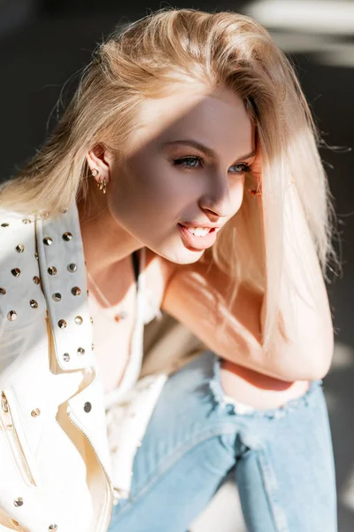 Retrato de uma mulher bonita feliz com belos olhos azuis com cabelo loiro com lábios sexy gordos em uma jaqueta de couro elegante primavera em um dia ensolarado. Bonito modelo menina goza de luz solar brilhante . — Fotografia de Stock