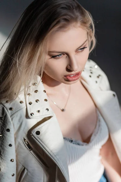 Retrato de uma bela jovem mulher com cabelo loiro com lábios sexy em uma camiseta elegante em uma jaqueta de couro elegante com rebites de metal em um dia ensolarado. Menina bonita olha para baixo e gosta do sol . — Fotografia de Stock