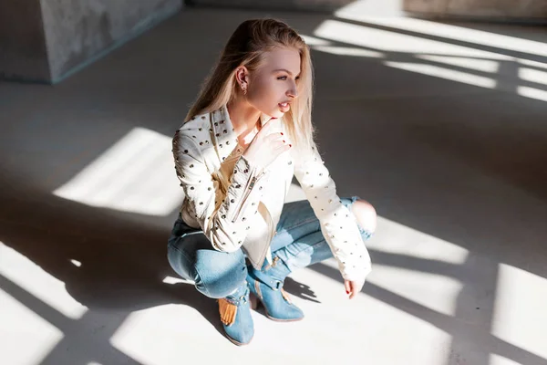 Europejska młoda kobieta model w stylu Vintage Kurtka skórzana w niebieskim modne zgrywanie dżinsy w zielonych modne buty kowbojskie pozowanie siedzi w pomieszczeniu ze światłem słonecznym. Zmysłowa stylowa dziewczyna relaksuje się w słonecznym studiu. — Zdjęcie stockowe