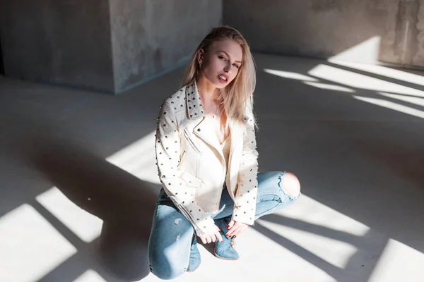 Glamorous modern attractive blond woman in vintage green cowboy boots in a summer leather jacket in ripped jeans sits in a room with concrete walls. Pretty beautiful girl model relaxes in the sun. — 图库照片