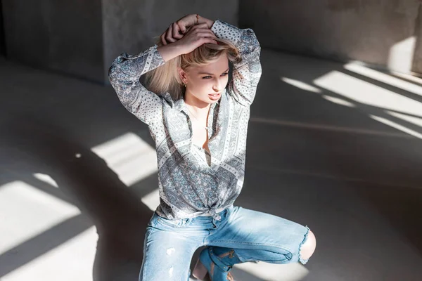 Stylowa seksowna młoda blond kobieta w rocznika letniej koszuli w modnej niebieskie dżinsy zgrywanie w zielonych buty kowbojskie pozowanie siedzi w pomieszczeniu ze światłem słonecznym. Całkiem piękna moda model dziewczyna cieszy się słońcem — Zdjęcie stockowe