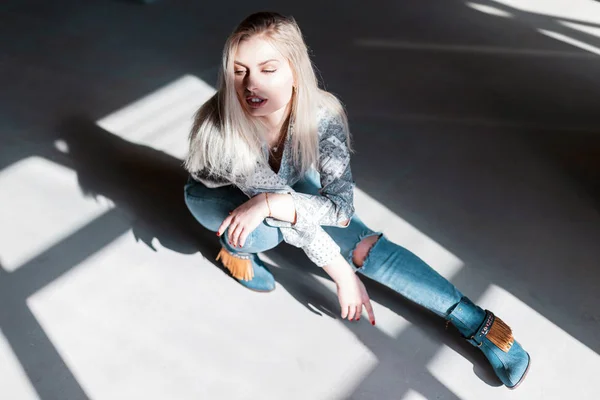 Moderne belle jeune femme modèle dans une chemise vintage avec un motif en bleu déchiré jeans en chaussures vertes posant dans un studio gris par une journée ensoleillée. Jolie fille blonde se détend au soleil. Bonne journée. . — Photo