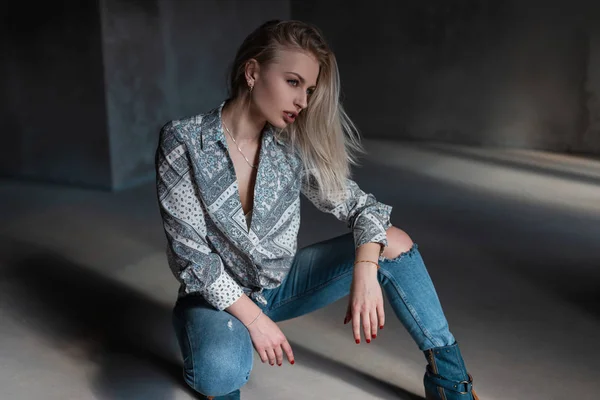 Bastante sexy hermosa modelo de mujer joven en una camisa vintage con un patrón en vaqueros rotos en zapatos elegantes posando en un estudio gris en un día soleado. Chica rubia atractiva está descansando. Ropa de estilo juvenil —  Fotos de Stock