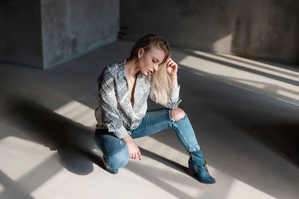 Bastante moderna jovem loira em camisa de verão elegante em azul rasgado jeans em botas de cowboy na moda posando sentado dentro de casa com luz solar. American bela moda modelo menina goza da luz do sol . — Fotografia de Stock