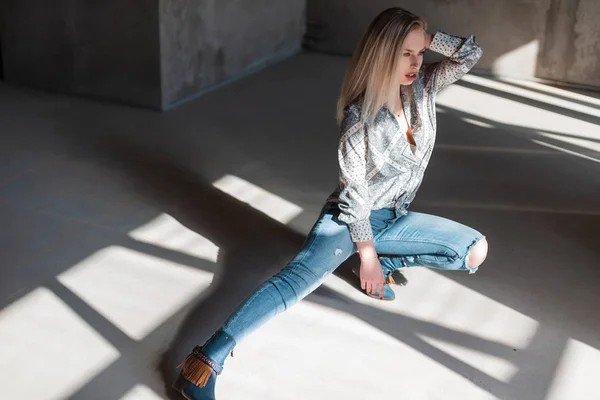 Amerykańska Młoda Blondynka w stylowej koszulce w Vintage zgrywanie dżinsy w modne buty kowbojskie pozowanie siedzi w pomieszczeniu ze światłem słonecznym. Seksowna piękna moda model dziewczyna relaksuje w słonecznym pomieszczeniu. — Zdjęcie stockowe