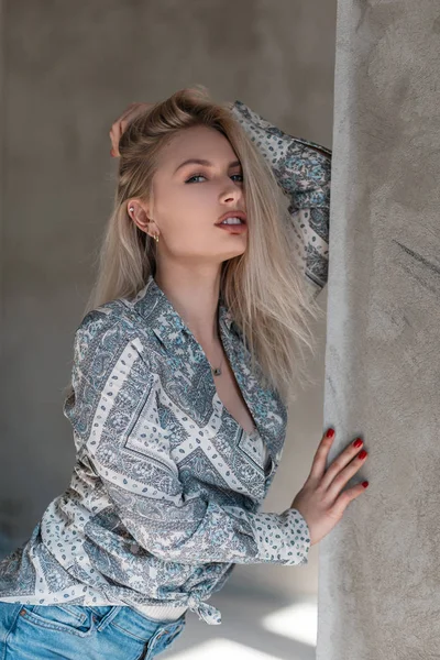 Красивая довольно сексуальная молодая блондинка в элегантной летней рубашке с узором в стильных джинсах стоит возле серой бетонной стены в помещении в солнечный день. Красивая девушка наслаждается выходными . — стоковое фото