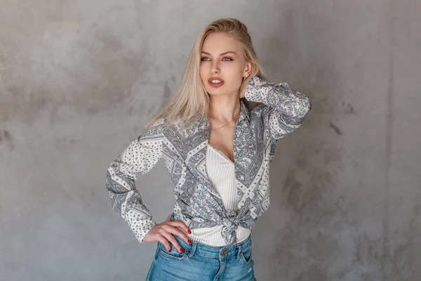 Sexy aantrekkelijke jonge vrouw een blonde in een modieus shirt met een patroon in een trendy t-shirt in stijlvolle jeans in de studio in de buurt van de Vintage grijze muur. Modern Europees meisje model. Jeugd stijl. — Stockfoto