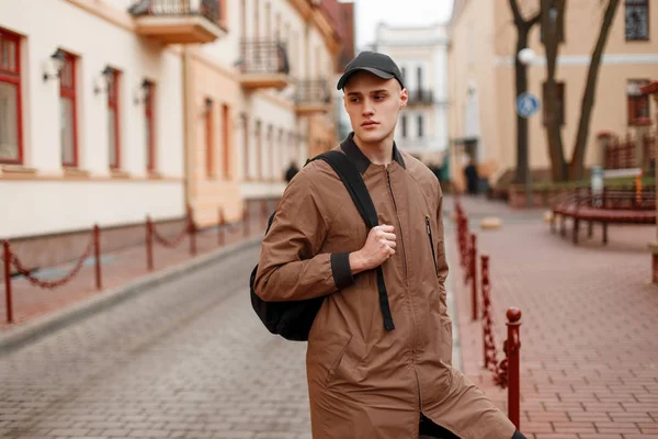 Amerikaanse modieuze knappe jonge man in een stijlvolle beige lange jas in een stijlvolle pet met een zwarte rugzak wandelingen door de stad op een lente dag. Aantrekkelijke vent buitenshuis. Spring Style herenkleding. — Stockfoto