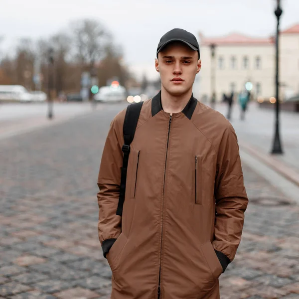 Stijlvolle knappe jonge man in een vintage beige lange jas in een zwarte modieuze pet met een zwarte rugzak reist rond de stad in de lente. Amerikaanse moderne man voor een wandeling. — Stockfoto