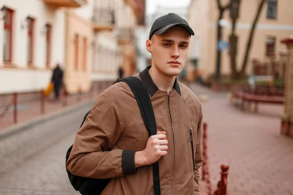 Portret van een jonge stijlvolle vent in een modieuze lente beige jas in een stijlvolle zwarte pet met een sportieve trendy rugzak in de stad. Amerikaanse knappe vent wandelingen in de straat. Mode. — Stockfoto