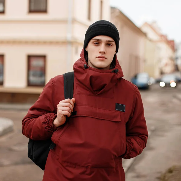 Fiatal jóképű magabiztos ember egy fekete kötött kalapot egy trendi piros hosszú kabát, fekete hátizsák áll a szabadban, közel szüreti épületeket. Szép modell fickó modellt sétálni. Őszi stílus. — Stock Fotó