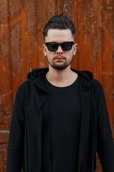 Serious brutal handsome young male hipster with fashionable hairstyle with beard in stylish sunglasses in modern black clothes is standing near the vintage wooden door to the bar. American guy. — Stock Photo, Image