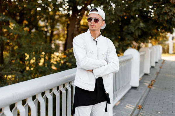 Jonge knappe Amerikaanse hipster man in een stijlvolle witte jas met een zonnebril in een modieuze zwarte t-shirt in trendy jeans poseert in een park op een zonnige zomerdag. — Stockfoto