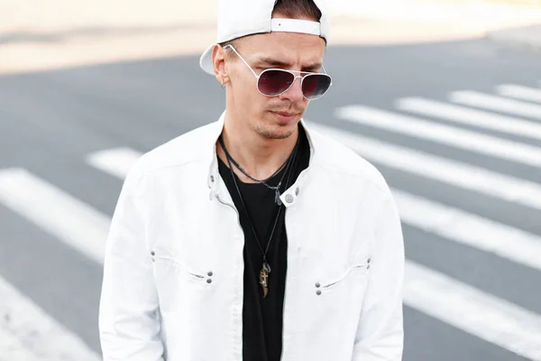 Moderne beau jeune homme hipster dans des lunettes rondes à la mode dans une veste blanche d'été dans une casquette de baseball à la mode dans un t-shirt noir se tient sur la route dans la ville. Un Américain dehors. Mode masculine . — Photo