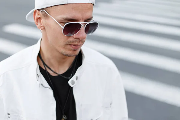 Retrato de close-up de um jovem hipster em uma jaqueta branca na moda em uma camiseta preta em um boné de beisebol elegante em óculos redondos na rua. Bonito cara moderno em um dia de verão . — Fotografia de Stock