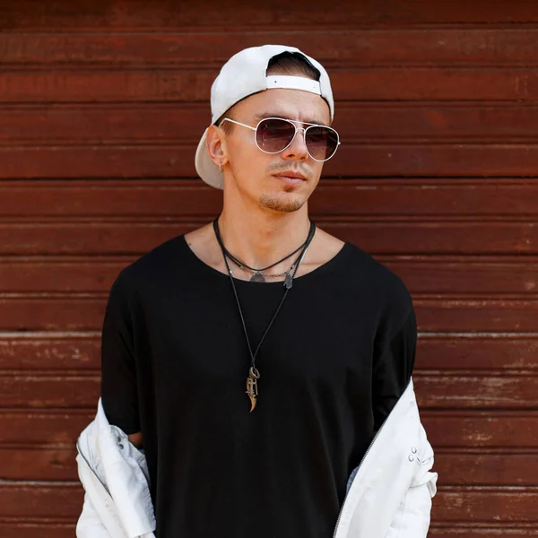 American handsome hipster fashionable man in dark sunglasses in a stylish white baseball cap in a black t-shirt in summer jacket with amulets around his neck near a vintage wooden wall. Cool guy. — Stockfoto