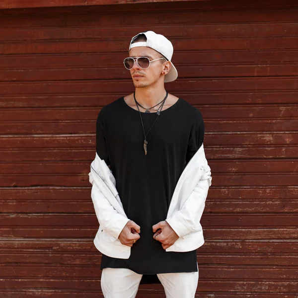 Hombre joven hipster guapo con una chaqueta de verano blanca de moda en una elegante camiseta negra con una gorra de béisbol de moda en gafas de sol oscuras cerca de una pared vintage de madera. Moda urbana con estilo guy model.Men — Foto de Stock