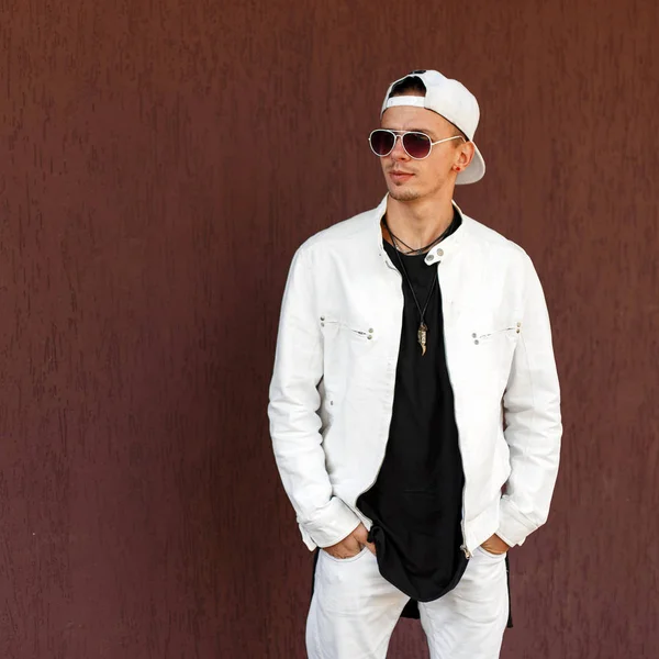 Nice hipster young man in a trendy white summer jacket in a stylish black t-shirt in a fashionable baseball cap in dark sunglasses near a vintage wall. Handsome modern guy model. Street style. — Stock Photo, Image