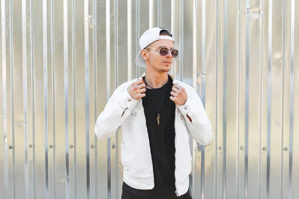 Handsome young hipster fashionable man in dark sunglasses in a stylish white jacket in a trendy baseball cap in a black t-shirt near a gray metal wall on a summer day. American city guy. — Stock Photo, Image