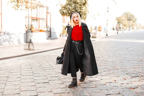 Mulher bonita bonita na moda em um casaco na moda preto longo com uma boina com uma bolsa de mão em sapatos elegantes em calças na moda em uma camiseta vermelha anda pela cidade em um dia ensolarado de outono. Menina bonito — Fotografia de Stock