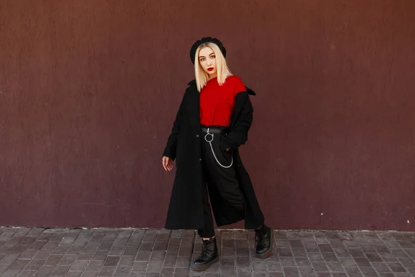 Hermosa mujer joven y elegante en un abrigo negro de moda con una camisa roja y zapatos de cuero negro de pie y posando cerca de la pared del color de pantone. Chica de moda de calle con labios rojos — Foto de Stock