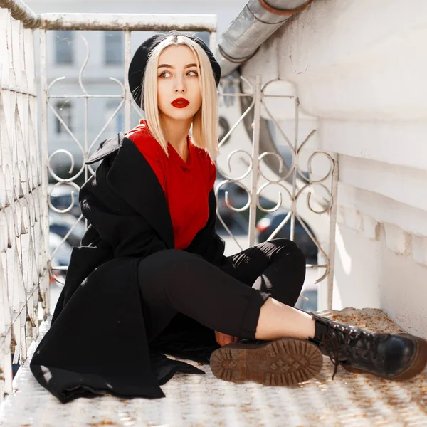 Jonge stijlvolle blonde vrouw met rode lippen in een modieuze jas met een zwarte baret in een elegante rode blouse in lederen laarzen zit in de buurt van een vintage metalen leuning op een zonnige herfst dag. Sexy meid. — Stockfoto