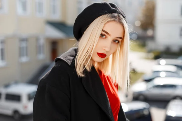 Portret van een glamoureuze mooie blonde vrouw met rode sexy lippen in een zwarte vacht in een elegante baret in een stijlvol rood shirt op een zonnige warme dag. Aantrekkelijk meisje fashion model geniet van de zon. — Stockfoto