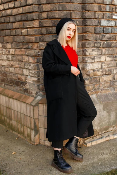 가죽 부츠에 빨간 셔츠에 검은 베 레모에 세련 된 롱 코트에서 금발 우아한 매력적인 젊은 여자는 도시에서 빈티지 벽돌 벽 근처에 서 있다. 귀여운 소녀 모델에 a 도보. — 스톡 사진