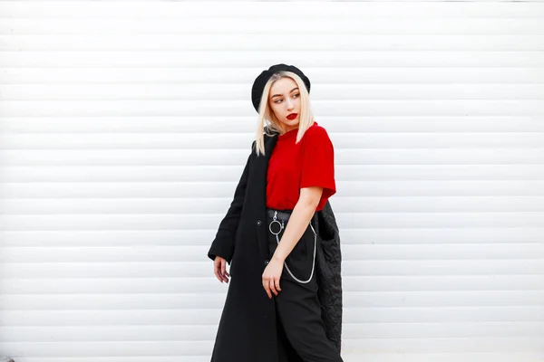 Beautiful pretty stylish young woman in an elegant beret in a black coat in a red shirt with red sexy lips in fashionable black pants near a modern metal wall outdoors. Pretty girl blond. Retro style. — ストック写真