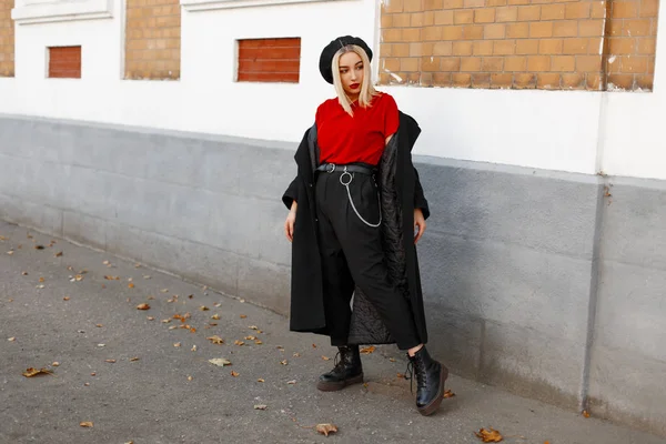 Mujer rubia linda de moda con un elegante abrigo negro en pantalones negros con una camiseta roja en una boina en botas de cuero está descansando de pie cerca del edificio en un día de otoño. Hermosa chica en la calle . — Foto de Stock