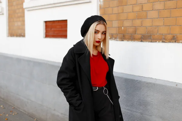 Glamorosa y hermosa joven moderna con una boina negra en una camiseta roja con un abrigo largo de moda en pantalones elegantes cerca de un edificio vintage en un día de otoño. Chica urbana de moda en un paseo . — Foto de Stock